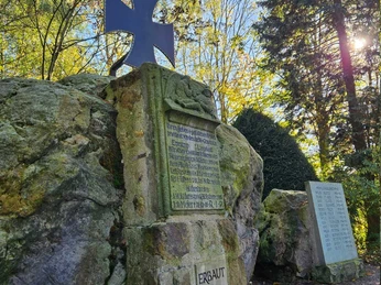 Erpentrup Ehrenmal Gedenkstein aus Naturstein mit schwarzem Kreuz und Namenstafeln, umgeben von Bäumen im Sonnenlicht.