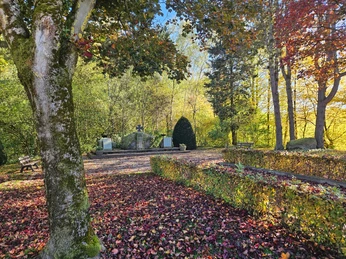 Ehrenmal inmitten herbstlich gefärbter Bäume, mit Steinmauer, Holzbänken und Laub auf dem Boden.