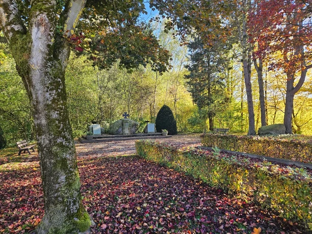 Ehrenmal inmitten herbstlich gefärbter Bäume, mit Steinmauer, Holzbänken und Laub auf dem Boden.
