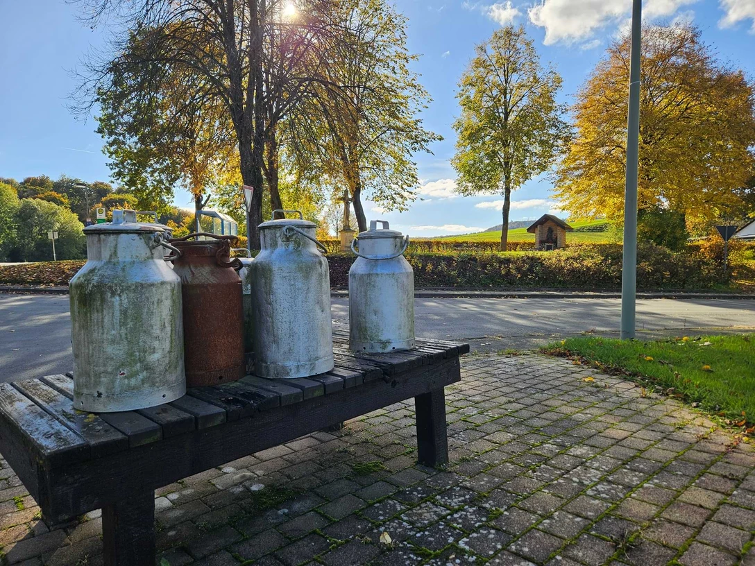 Mehrere alte Milchkannen auf einer Holzbank am Straßenrand vor herbstlicher Landschaft.