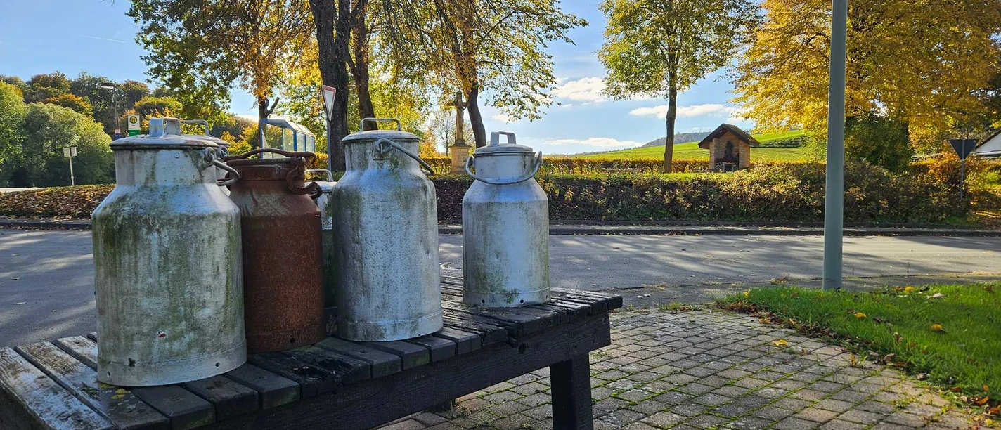 Erpentrup Milchkannen Mehrere alte Milchkannen auf einer Holzbank am Straßenrand vor herbstlicher Landschaft.