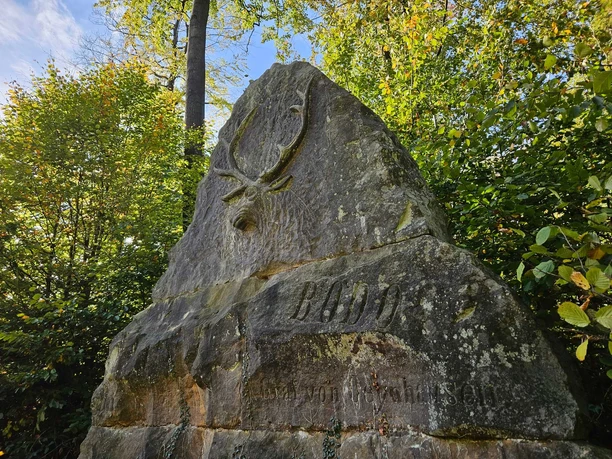 Großer Gedenkstein mit Hirschrelief und Inschrift „BODO“, umgeben von grünem Laubwald.