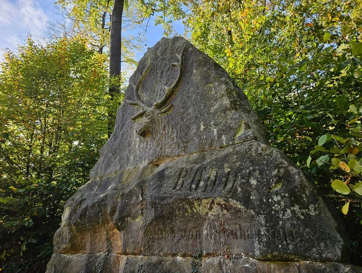Großer Gedenkstein mit Hirschrelief und Inschrift „BODO“, umgeben von grünem Laubwald.