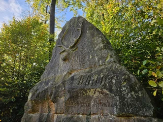 Langeland Bodostein Großer Gedenkstein mit Hirschrelief und Inschrift „BODO“, umgeben von grünem Laubwald.