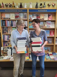 HelgaKatja webversion.jpg Two women stand in front of bookshelves labeled 'The special book' and 'Bestseller', each holding a large stack of books.