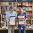 HelgaKatja webversion.jpg Zwei Frauen stehen vor Bücherregalen mit den Überschriften 'Das besondere Buch' und 'Bestseller' und halten jeweils einen großen Stapel Bücher.