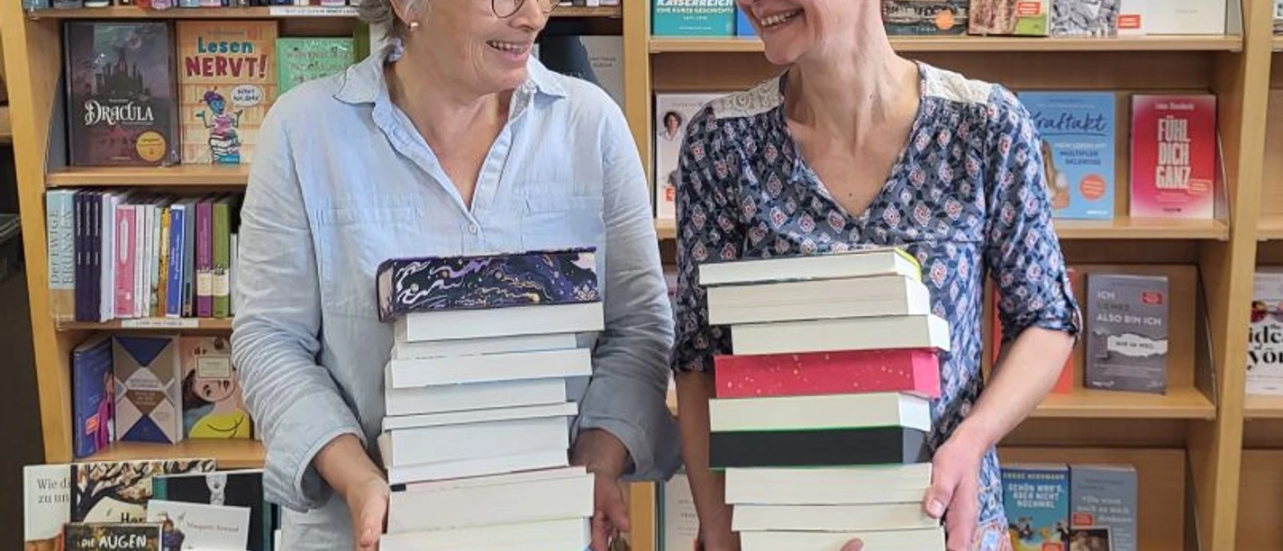 HelgaKatja webversion.jpg Two women stand in front of bookshelves labeled 'The special book' and 'Bestseller', each holding a large stack of books.