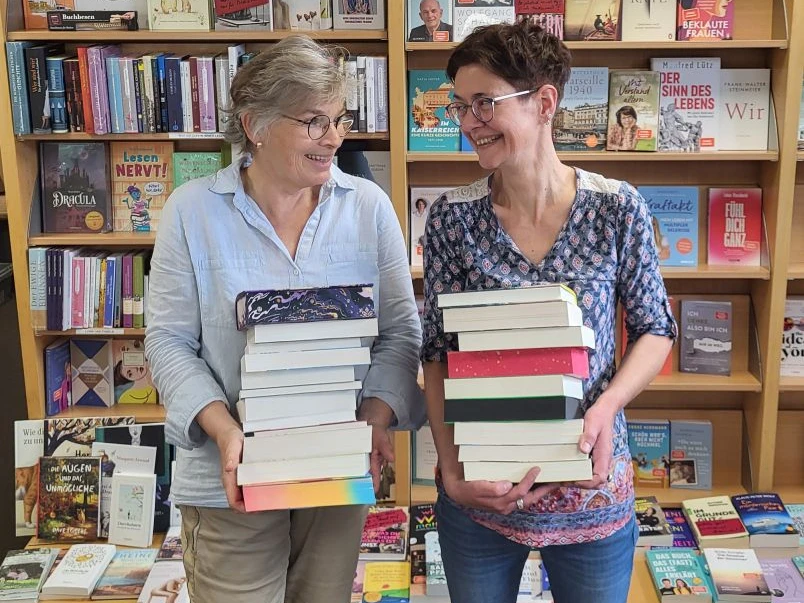 HelgaKatja webversion.jpg Zwei Frauen stehen vor Bücherregalen mit den Überschriften 'Das besondere Buch' und 'Bestseller' und halten jeweils einen großen Stapel Bücher.