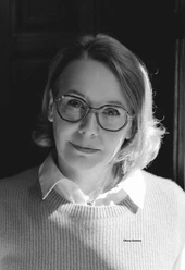 Sandra Lüpkes Webversion.jpg Person with shoulder-length, wavy hair, white shirt and light gray sweater against a dark background.