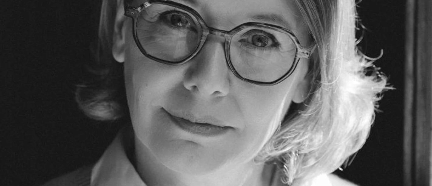 Sandra Lüpkes Webversion.jpg Person mit schulterlangem, gewelltem Haar, weißem Hemd und hellgrauem Pullover vor dunklem Hintergrund.