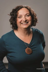Oswald Susanne Webversion.jpg Person with shoulder-length curly hair wears a dark green top and a round wooden necklace.
