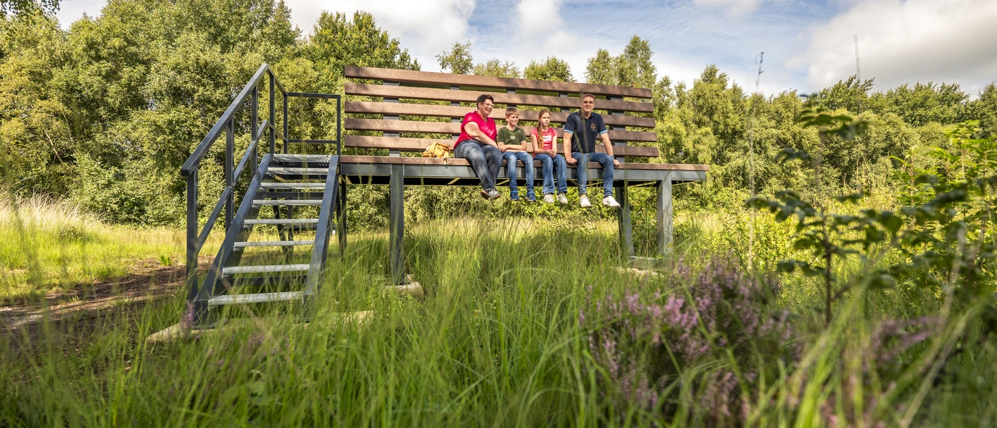Familienausflug - XXL-Bank im Provinzialmoor ©Emsland Tourismus GmbH (2) Familie sitzt auf einer großen Bank im Moor, umgeben von grüner Wiese und Bäumen unter blauem Himmel.