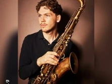 Ein junger Mann hält ein Saxophon in dunkler Umgebung.