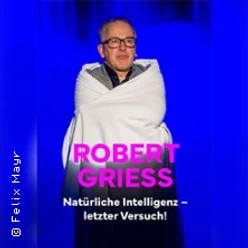 Robert Griess auf einer Bühne, in eine Decke gehüllt, vor blauem Hintergrund.