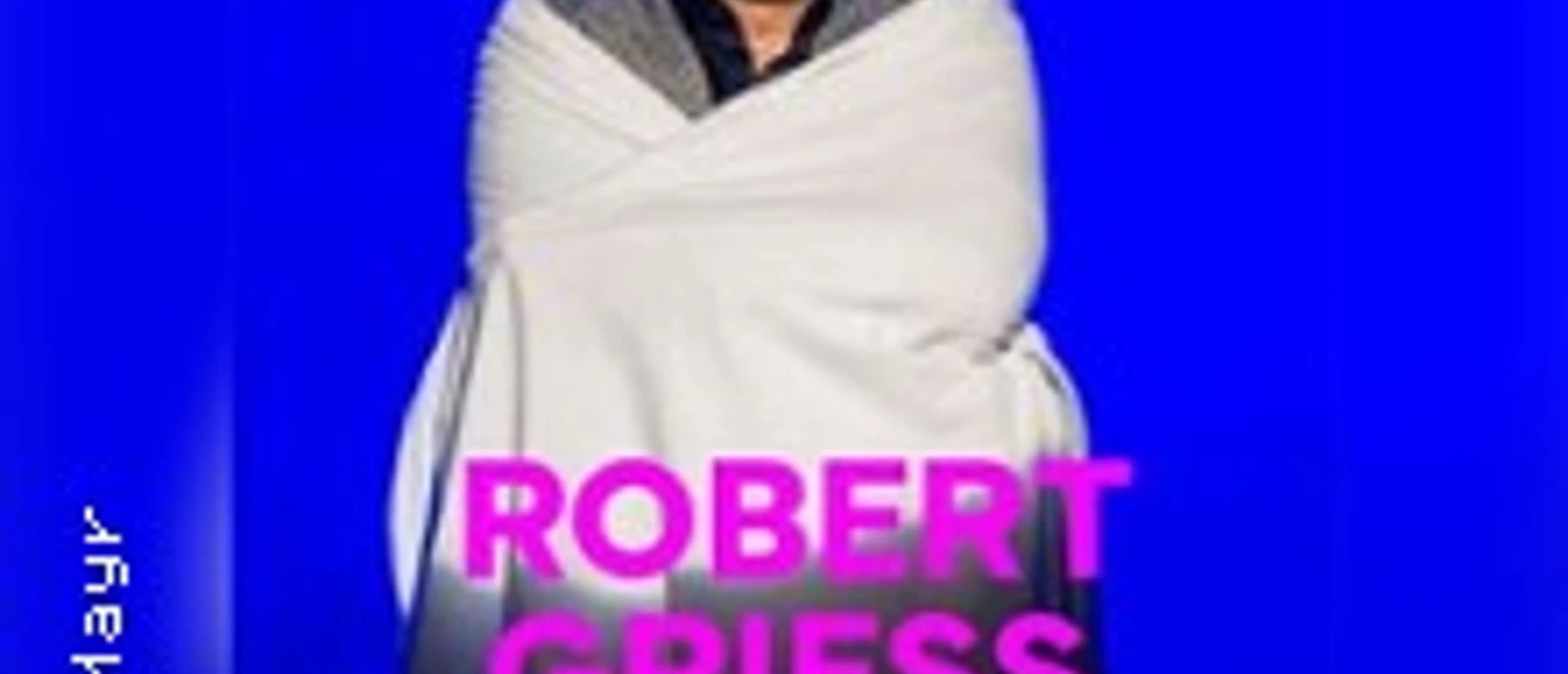 Robert Griess auf einer Bühne, in eine Decke gehüllt, vor blauem Hintergrund.