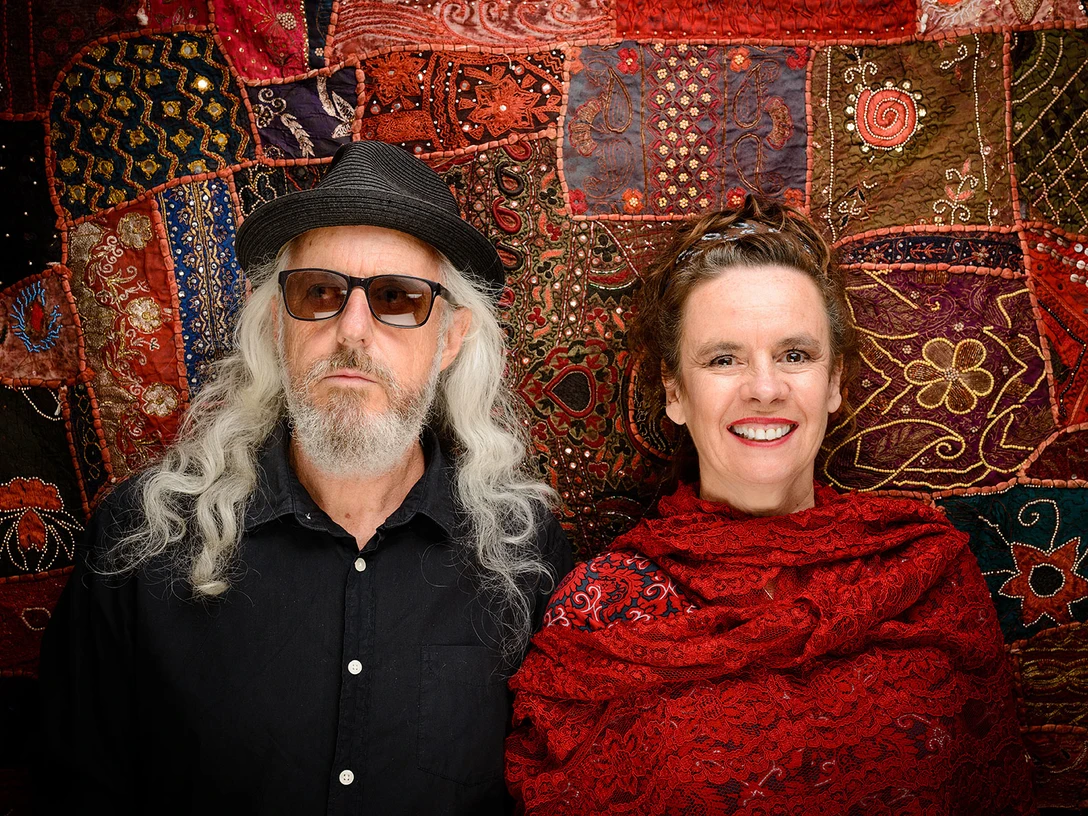 Nessi Tausendschön und William Mackenzie kommen nach Espelkamp Ein Mann mit grauem Haar und Sonnenbrille neben einer lachenden Frau in rotem Tuch vor buntem Stoff.