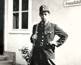 01 Heinrich Jansen_Querformat.jpg Historische Aufnahme eines uniformierten Grenzbeamten mit Gewehr vor einem Gebäude mit Schild.