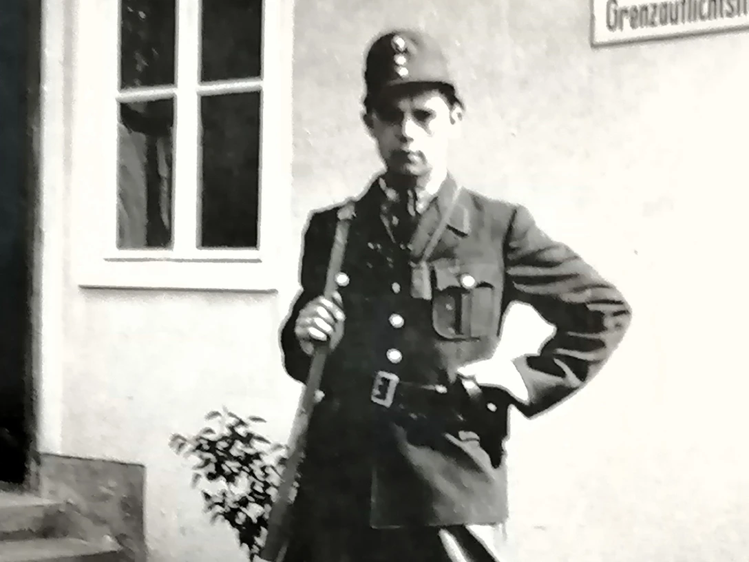 01 Heinrich Jansen_Querformat.jpg Historische Aufnahme eines uniformierten Grenzbeamten mit Gewehr vor einem Gebäude mit Schild.