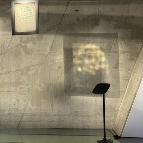 Lichtmotiv.jpeg Projiziertes Schwarzweiß-Porträt auf einer rauen Betonwand, daneben ein schwarzer Informationsständer unter einer Deckenleuchte.