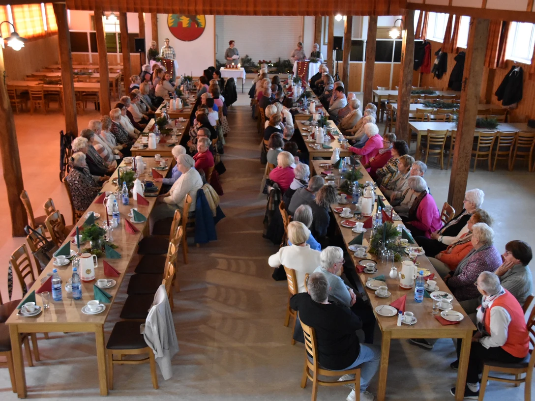 DSC_0016-3.JPG Menschen sitzen an langen gedeckten Holztischen in einer Halle und nehmen gemeinsam an einer Feier teil.