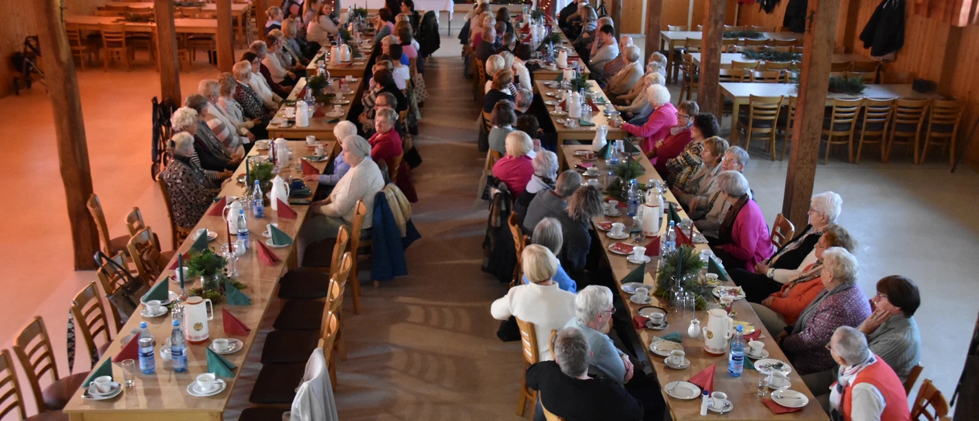 DSC_0016-3.JPG Menschen sitzen an langen gedeckten Holztischen in einer Halle und nehmen gemeinsam an einer Feier teil.