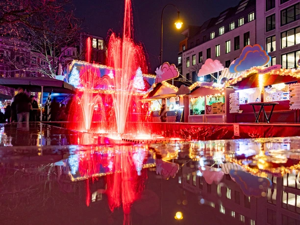 HEAVENUE Cologne A festively lit Christmas market with colorful stalls and a red fountain at night.
