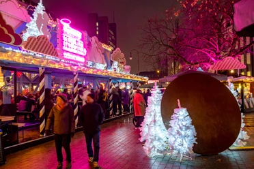 Bunt erleuchteter Weihnachtsmarkt mit leuchtenden Engeln und weißen Tannen in der Abenddämmerung.