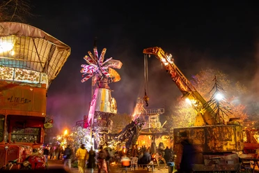 At night, imaginative sculptures and machines shine in colorful light, surrounded by people.