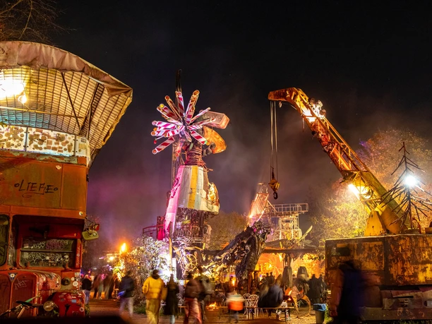 At night, imaginative sculptures and machines shine in colorful light, surrounded by people.