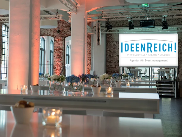 IdeenReich! GmbH - Veranstaltungsraum Veranstaltungsraum mit Backsteinwänden, weißen Säulen und Hochtischen.