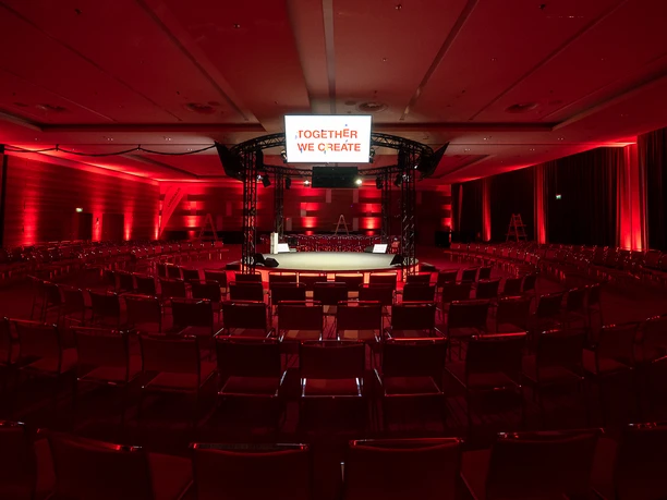 IdeenReich! GmbH - Event room with seating Event room bathed in red light with row seating and a brightly lit stage.