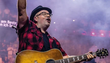 Björn Heuser Musician Björn Heuser wears black glasses and a hat as he sings with an acoustic guitar in his hand. His dynamic posture and open lips suggest a lively performance.