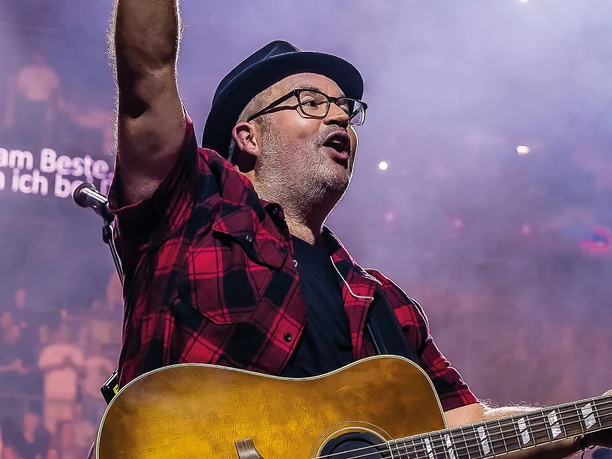 Björn Heuser Musician Björn Heuser wears black glasses and a hat as he sings with an acoustic guitar in his hand. His dynamic posture and open lips suggest a lively performance.