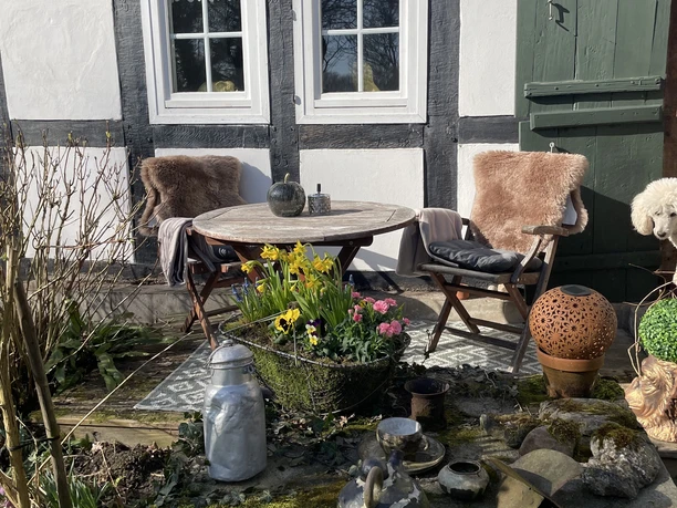 Ferienwohnung "wie bei Muddi" Sitzplatz vor Fachwerkhaus mit Holztisch, zwei Stühlen, Fellauflagen, Blumen und alten Gefäßen.