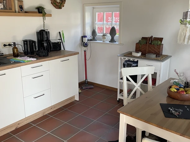 Ferienwohnung "wie bei Muddi" Kleine Küche mit Holztisch, weißen Stühlen, heller Arbeitsfläche und Küchenutensilien auf Fliesenboden.