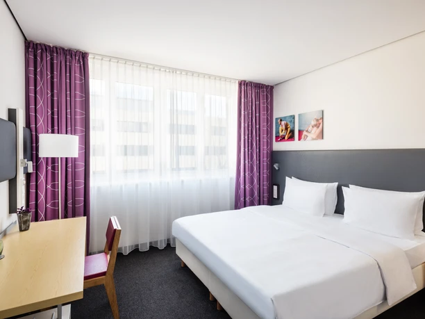 Lindner Hotel Frankfurt Sportpark - Hotel rooms Bright hotel room with double bed, desk and purple curtains