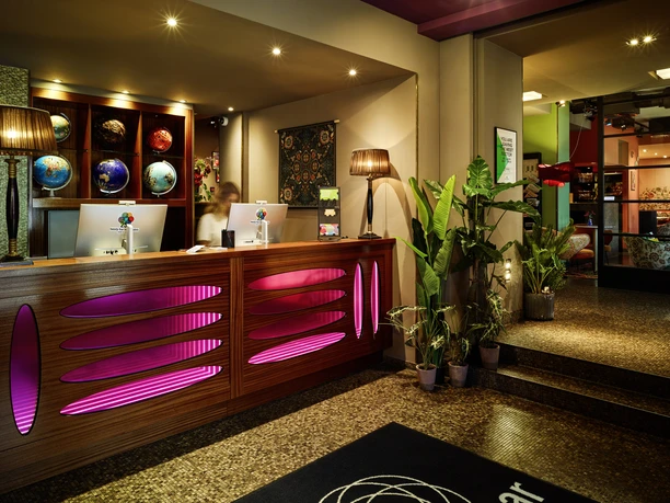25hours Hotel the Goldman - Hotelrezeption Hotelrezeption mit pink beleuchtetem Tresen und einer Sammlung von Globen.