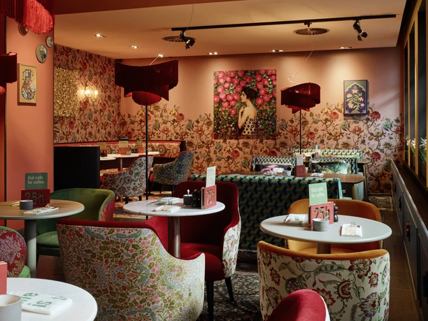 25hours Hotel the Goldman - Farbenfroher Restaurantbereich Farbenfroher Restaurantbereich mit floraler Tapete, rosa Wänden und bunten Sesseln.