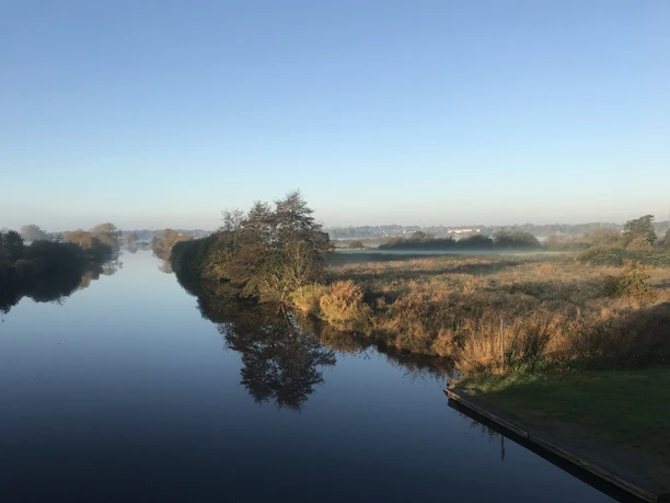Anlegestelle Stoteler Brücke Flusslauf mit Morgennebel, Spiegelung von Bäumen im stillen Wasser unter klarem blauem Himmel.