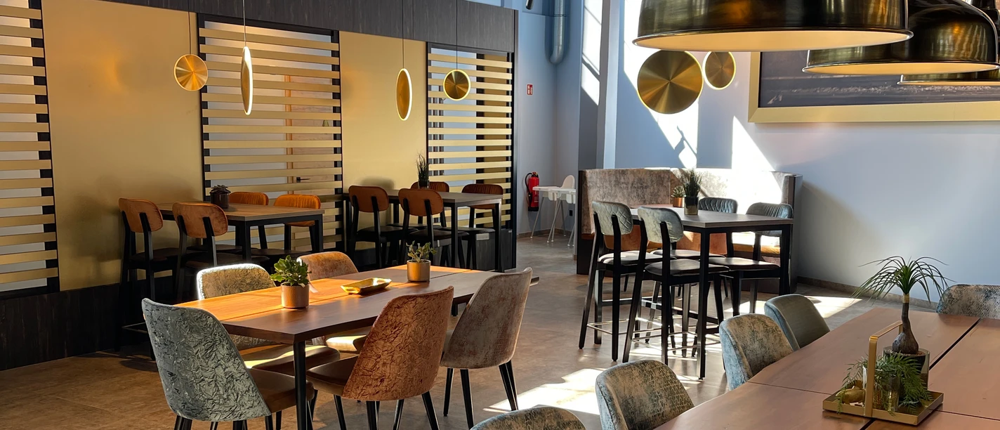 IMG_7300 (c) Kai-Uwe Schulz_Erlebnis Bremerhaven_Ausschnitt.jpg Helles Restaurant mit modernen Holztischen, Polsterstühlen und großen goldfarbenen Lampen.