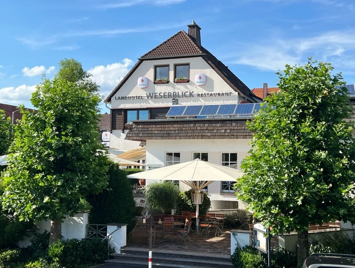 Fassade des Landhotels Weserblick mit Restaurantterrasse, Sonnenschirmen und grünen Bäumen.