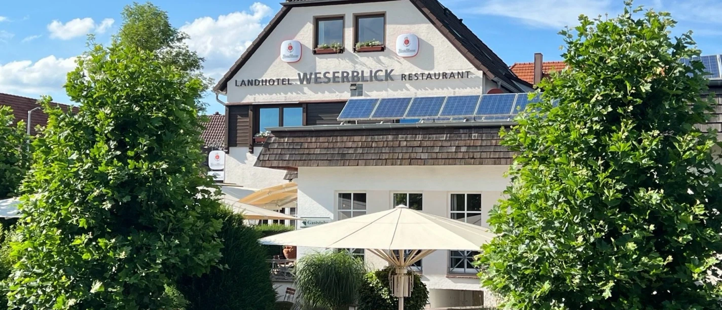 Landhotel Weserblick Fassade des Landhotels Weserblick mit Restaurantterrasse, Sonnenschirmen und grünen Bäumen.