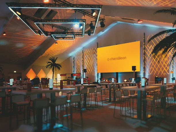 Chamäleon Location - Innenansicht Eventlocation mit gedämpftem Licht, hohen Palmen und Brückentischen..