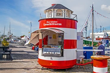 InstaWalk Fischereihafen (c) Michael Schmidt (14).jpg Rot-weißer Imbissstand in Leuchtturmform im Fischereihafen Bremerhaven mit Hafenbetrieb im Hintergrund.