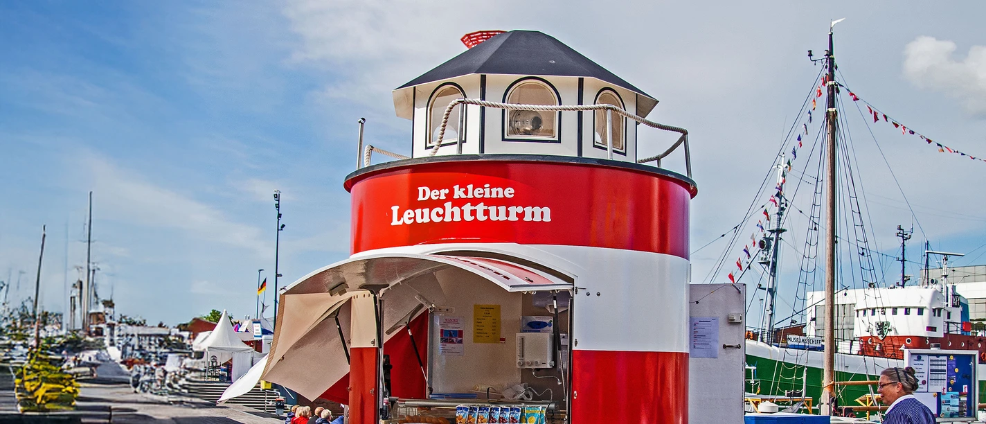 InstaWalk Fischereihafen (c) Michael Schmidt (14).jpg Rot-weißer Imbissstand in Leuchtturmform im Fischereihafen Bremerhaven mit Hafenbetrieb im Hintergrund.