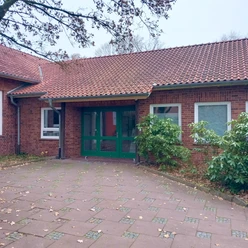 Backsteingebäude mit rotem Ziegeldach, begrüntem Eingang und gepflastertem Vorplatz.