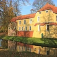 Manningaburg Pewsum Gelbes Backsteingebäude mit rotem Dach, umgeben von Wassergraben und herbstlich belaubten Bäumen.