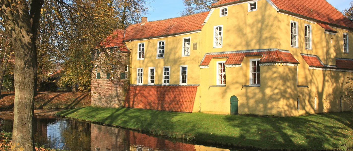 Manningaburg Pewsum Gelbes Backsteingebäude mit rotem Dach, umgeben von Wassergraben und herbstlich belaubten Bäumen.