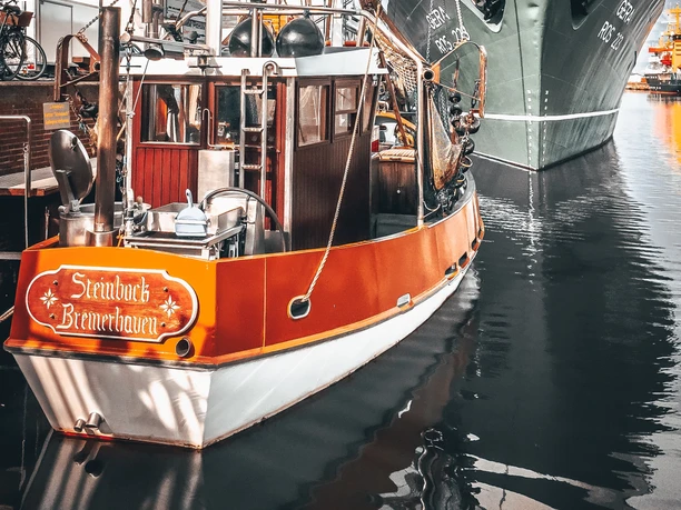 InstaWalk Fischreihafen (c) Sandra Chizzali (4).jpg Krabbenkutter Steinbock mit rotem Rumpf liegt im Fischereihafen neben einem großen Frachtschiff.