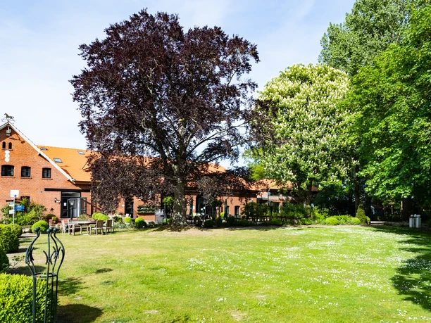 Gut Landegge_5107-HDR.jpg Backsteingebäude mit rotem Dach hinter grüner Wiese und blühenden Bäumen unter blauem Himmel.